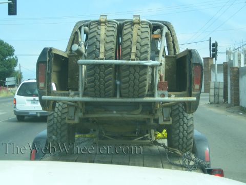Rear of a muddy off-road truck on a trailer carrying two spare tires on a highway