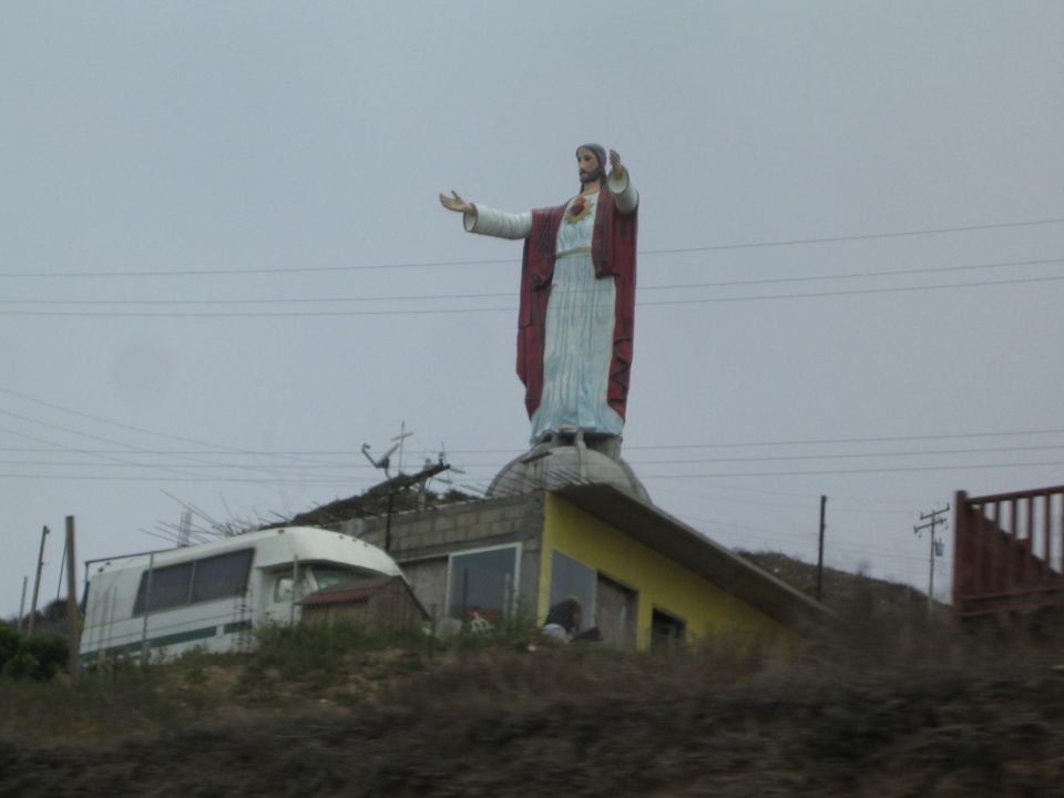 Roadside statue of Jesus with outstretched arms atop a small building on a hill