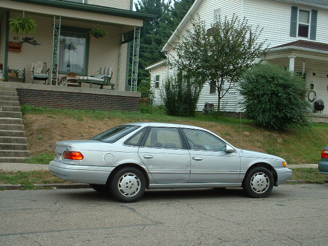 Silver Ford Taurus sedan parked curbside, viewed from the passenger side on a residential street