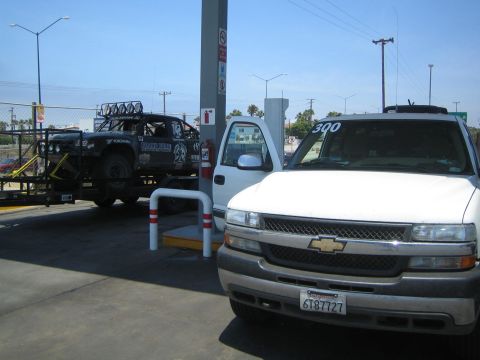 White Chevrolet truck at a gas station with a black race truck on a trailer behind