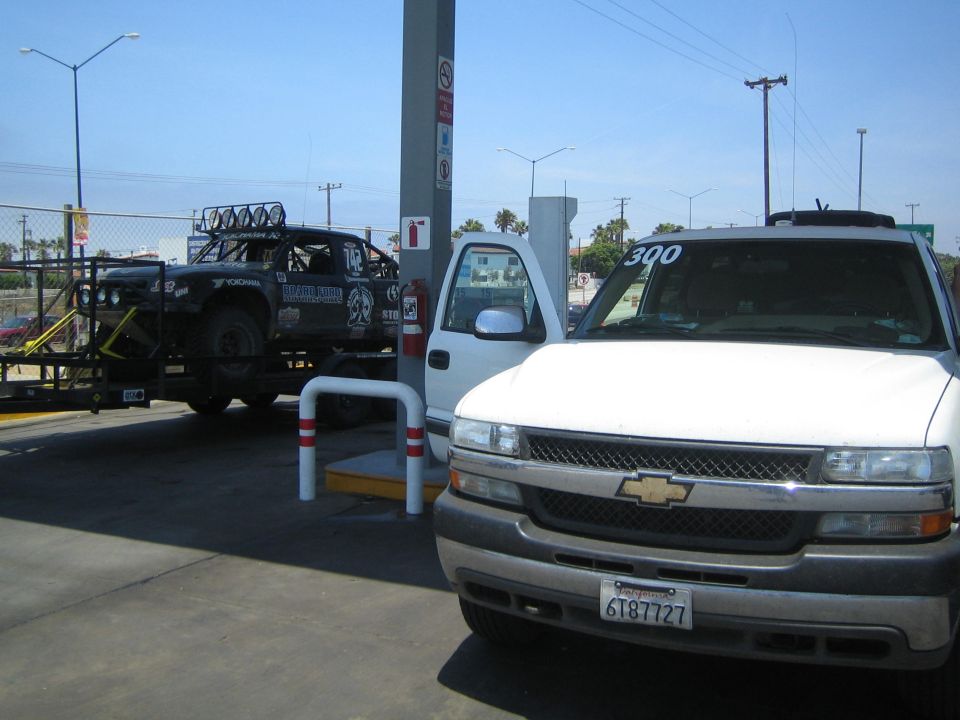 White Chevrolet truck at a gas station with a black race truck on a trailer behind