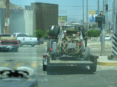 Tan off-road buggy number 946 strapped on a flatbed trailer in city traffic