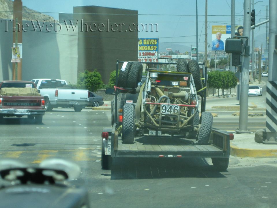 Tan off-road buggy number 946 strapped on a flatbed trailer in city traffic