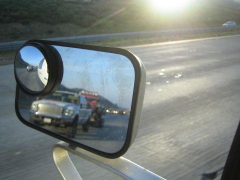 Side mirror reflecting a flatbed truck hauling an off-road vehicle at sunrise