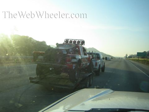 Red off-road race truck on a trailer towed by a white pickup at sunrise