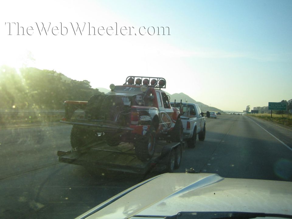 Red off-road race truck on a trailer towed by a white pickup at sunrise