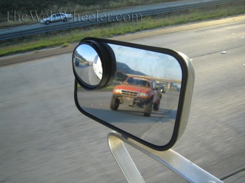Side mirror reflecting an orange off-road truck on a highway