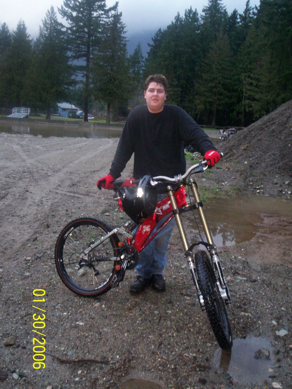 Man in black shirt and jeans with red gloves stands next to a red downhill mountain bike on a dirt path