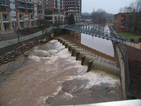 1978 Ford Bronco - A wide view of a river overflowing its banks, with a pedestrian bridge spanning across it and modern buildings lining the shore.