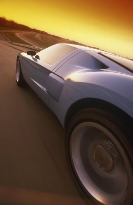 2003 Ford GT - A blue sports car is captured in motion from a rear quarter angle against a vibrant sunset sky.