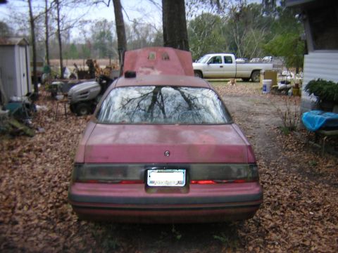 1987 Ford Thunderbird - The rear of a red 1987 Ford Thunderbird is visible, with its hood propped open and a white pickup truck in the background.