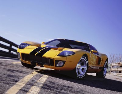 2003 Ford GT - A yellow 2005 Ford GT with black racing stripes is captured in a dynamic shot on a road.