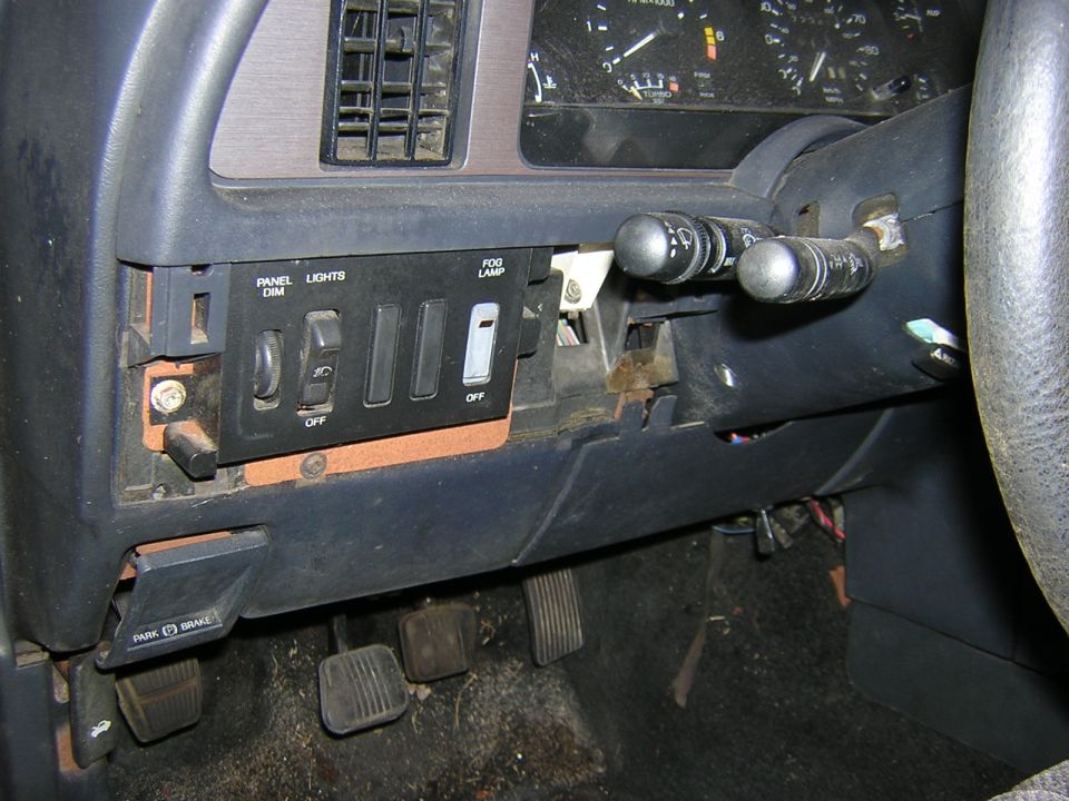 Car interior dashboard with panel lights, fog lamp switches, and pedals visible. The dashboard is dark gray