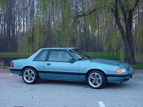 1994 Ford Bronco - Side view of a light blue coupe, likely a 1990 Ford Mustang, parked outdoors with trees in the background.