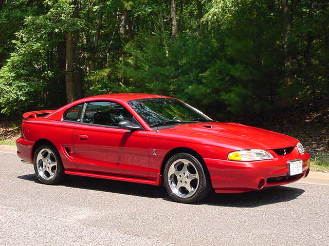 Red 1994 Ford Mustang parked on asphalt with trees in background. Supermotors.net logo visible