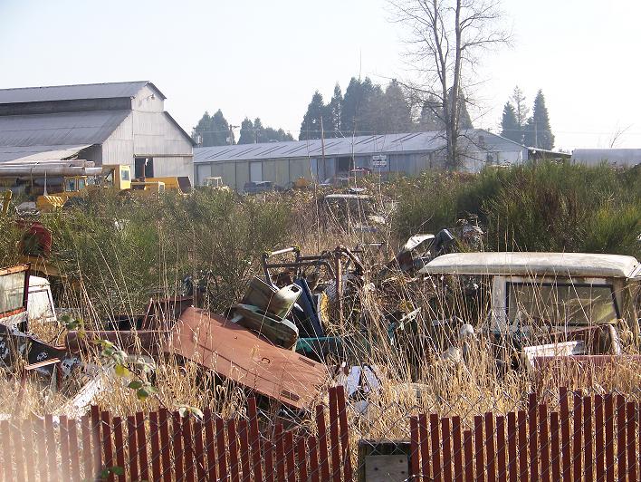 Junkyard scene with overgrown grass, various vehicle parts, and buildings in the background