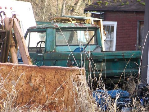1968 Ford Bronco - An old green pickup truck sits abandoned in overgrown weeds, partially obscured by a rusty metal container.