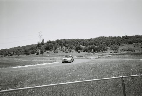 1988 Mazda MX-6 - A silver sedan navigates a turn on a race track, with grassy fields and trees in the background.