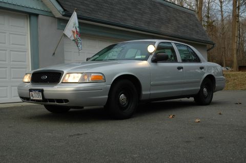 2003 Ford Crown Victoria - Silver 2003 Ford Crown Victoria police car with a spotlight on the driver's side is parked in front of a garage.