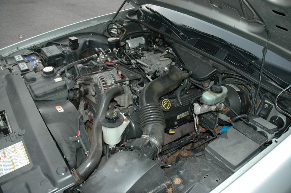 Engine bay of a silver Ford sedan showing a V8 with intake tube and fluid reservoirs