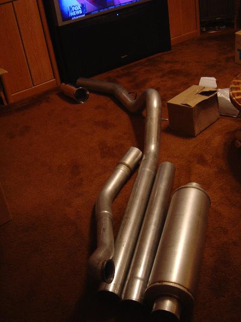 1986 Ford Bronco II - Various exhaust pipes and a muffler are laid out on a carpeted floor, with a television visible in the background.