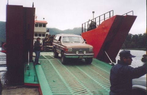 1989 Ford Bronco - A brown Ford Bronco is being driven onto a ferry, with a large red structure and another vessel in the background.