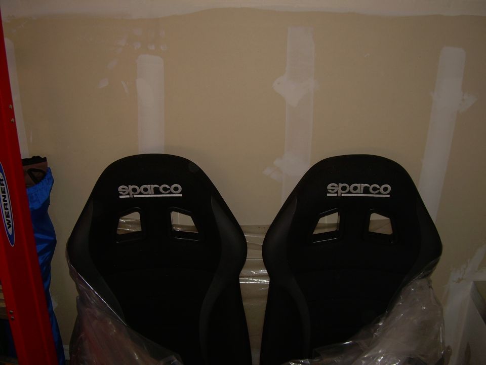 Two black Sparco racing seats with white logos, partially wrapped in plastic