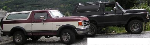 1989 Ford Bronco - Two Ford Broncos, a two-tone maroon and white model and a black one, are parked side-by-side.