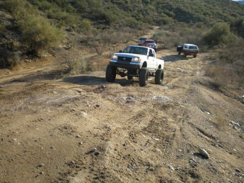 1991 Ford Bronco - Several trucks navigate a dusty, off-road trail in a hilly, arid landscape.