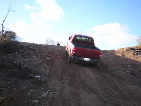 1991 Ford Bronco - A red pickup truck navigates a steep dirt incline under a partly cloudy sky.