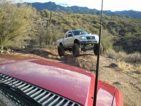1991 Ford Bronco - A lifted silver Toyota Tacoma navigates a desert trail, with another vehicle's hood in the foreground.