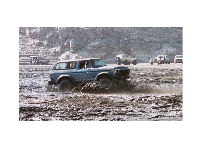 Blue Ford Bronco blasting through a muddy pit with other trucks in the distance