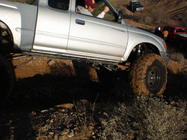 Silver pickup truck with large, mud-covered tires driving on a dirt trail. Another red truck and a dark SUV are visible in the background