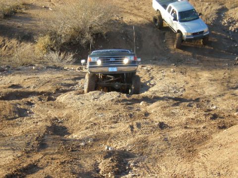 1991 Ford Bronco - Two trucks navigate a challenging off-road terrain with steep inclines and loose dirt.