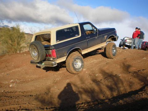 1991 Ford Bronco - A dark blue and tan Ford Bronco navigates a dirt incline with other vehicles and people in the background.