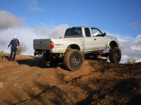 1991 Ford Bronco - Silver Toyota Tacoma TRD truck with oversized tires navigates a dirt incline under a blue sky with scattered clouds.