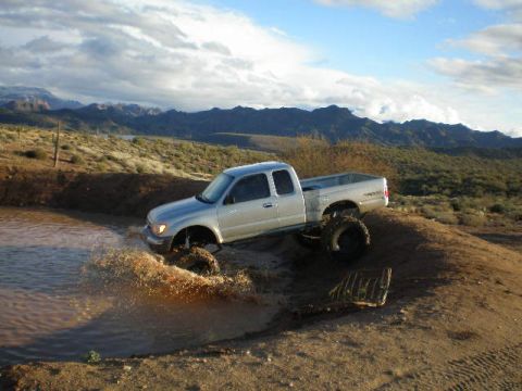 1991 Ford Bronco - A silver pickup truck navigates a muddy water obstacle in a desert landscape.