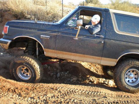 1991 Ford Bronco - A driver steers a dark blue 1991 Ford Bronco with a lifted suspension through a challenging off-road terrain.