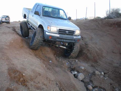 1991 Ford Bronco - A modified Toyota Tacoma with large off-road tires tackles a challenging dirt trail, with another vehicle in the distance.