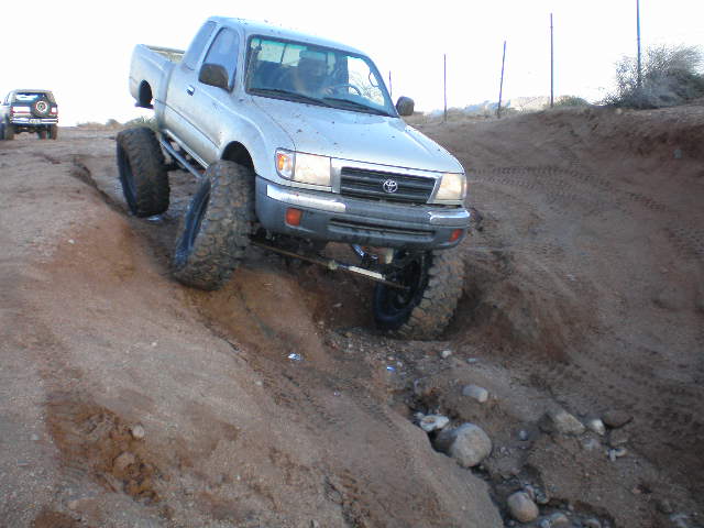 Silver Toyota Tacoma with oversized tires navigating a steep dirt incline; another SUV is visible in the background