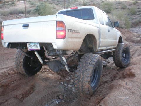 1991 Ford Bronco - Modified Toyota Tacoma pickup truck with off-road tires and suspension lift driving on dirt.