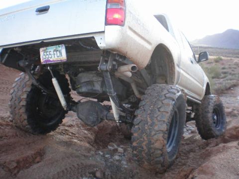 1991 Ford Bronco - This lifted pickup truck is navigating a rough, dirt-covered terrain with large, knobby tires.