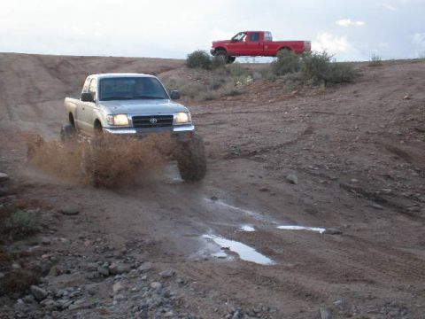 1991 Ford Bronco - A silver Toyota Tacoma navigates a muddy off-road trail, with a red pickup truck visible on a distant hill.