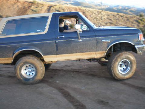 1991 Ford Bronco - A lifted 1991 Ford Bronco is seen driving on a dirt road in a dry, hilly landscape.