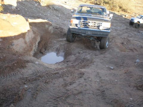 1991 Ford Bronco - A dark blue pickup truck navigates a rough dirt path with a puddle and another vehicle visible.
