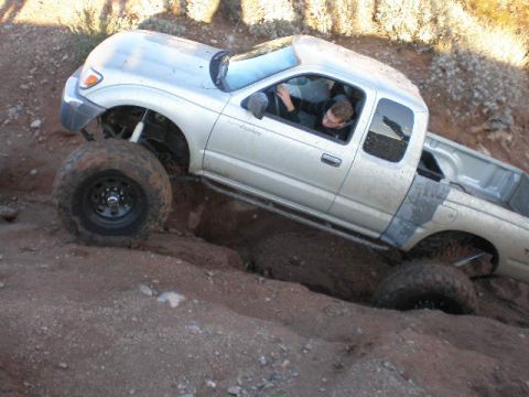 1991 Ford Bronco - A silver pickup truck is shown off-roading on a dirt trail, with one wheel significantly elevated.