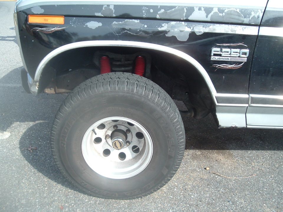 Front wheel of black lifted Ford with F-250 badge, red coil springs, locking hub, Cooper tire, peeling paint