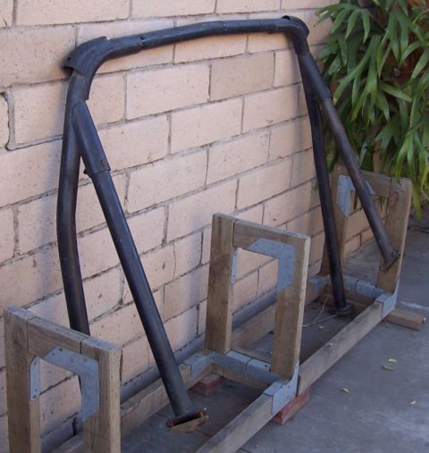 1990 Ford Bronco - A black roll bar is shown mounted on wooden supports, leaning against a brick wall with greenery in the background.