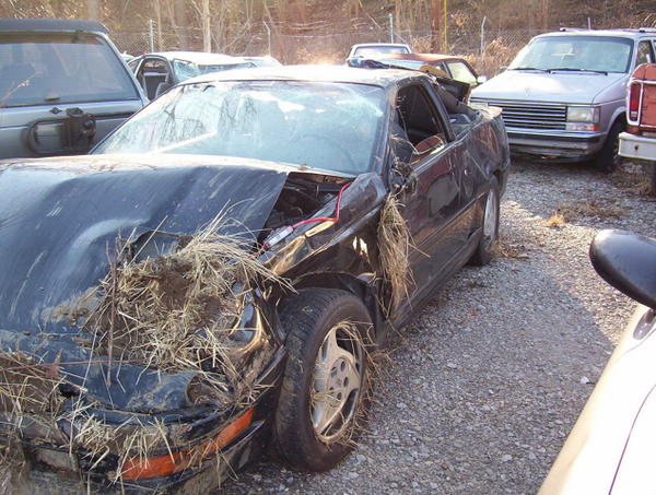 1995 Ford Probe with heavily damaged front end, grass and debris in engine bay, parked in a junkyard with other vehicles