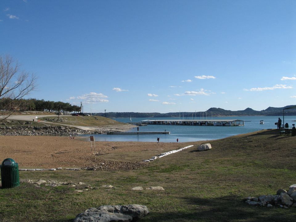 Lakeside beach with marina docks, sailboats and distant hills under a clear blue sky